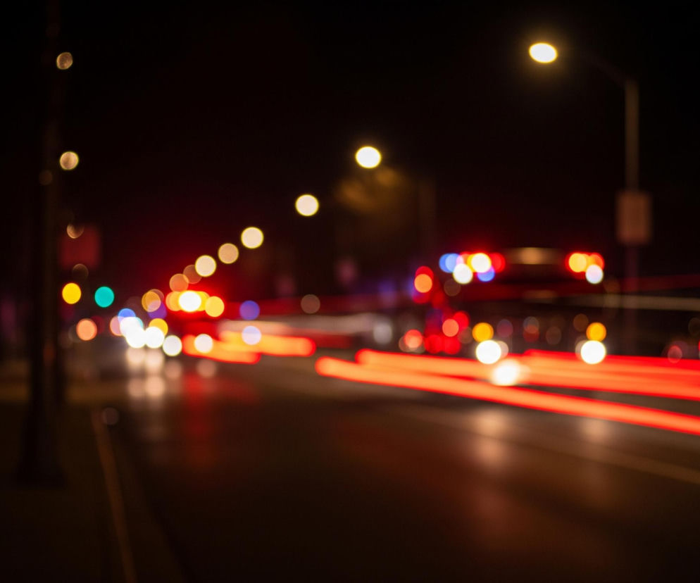Uliczna scena nocna, rozmyta i wypełniona światłami bokeh. Po prawej stronie widać rozmyte, poziome smugi czerwonych świateł, sugerujące ruch pojazdów, a także pojedyncze, okrągłe światła w kolorach białym, czerwonym i niebieskim. Na horyzoncie, po lewej, znajdują się smugi fioletowego i czerwonego światła, natomiast na prawym skraju, częściowo widoczny jest budynek o ciemnych oknach. Wzdłuż ulicy rozmieszczone są latarnie uliczne emitujące ciepłe, żółte światło, a na pierwszym planie, po lewej stronie, przebiega fragment chodnika.