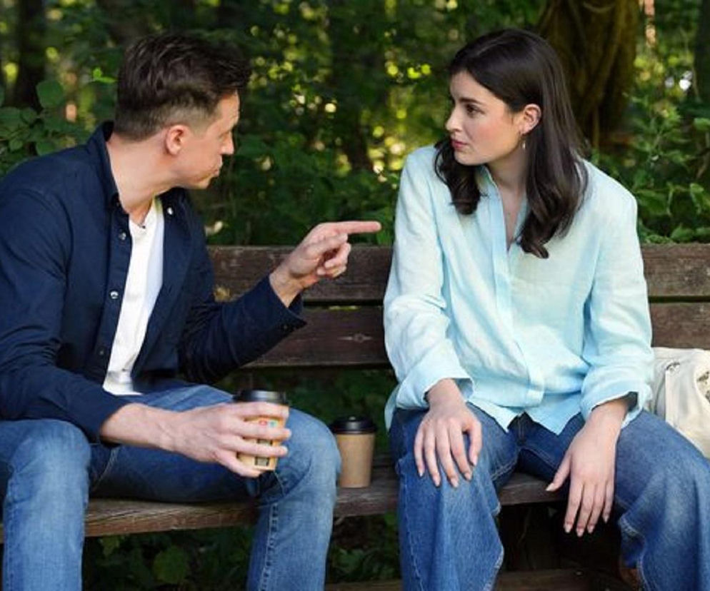 Błażej (Piotr Ligienza) i Weronika (Maria Świłpa) z serialu Barwy szczęścia siedzą na ławce w parku. Błażej, ubrany w granatową koszulę i dżinsy, trzyma kubek kawy i wskazuje palcem w stronę Weroniki. Ona, w błękitnej koszuli i dżinsach, z torebką obok, słucha go z wyraźnym niezadowoleniem. Więcej o ich relacji przeczytasz na Super Seriale.