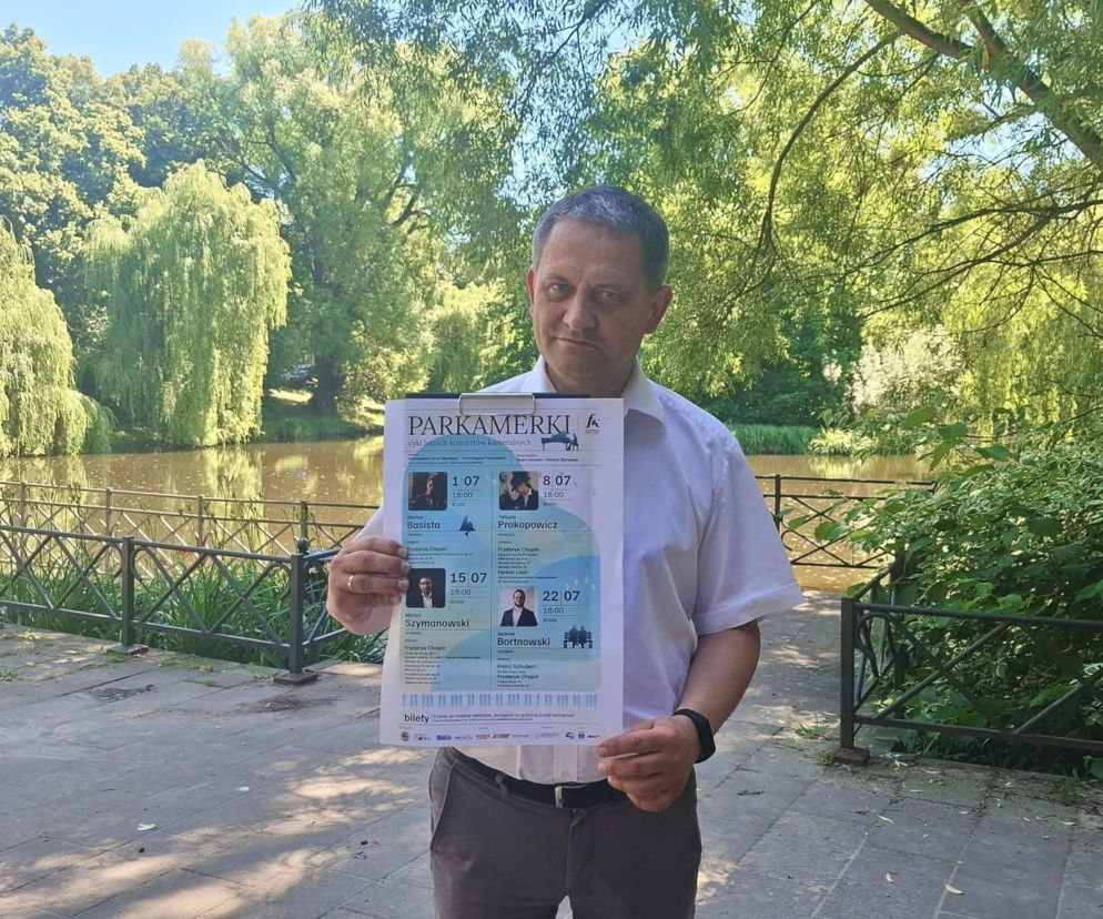 Mężczyzna w jasnej koszuli trzyma plakat informujący o cyklu koncertów „Parkamerki” w Koszalinie. Tłem jest malowniczy park z jeziorem i zieloną roślinnością. Na naszym portalu możesz przeczytać o tym wydarzeniu, które promuje muzykę Fryderyka Chopina.