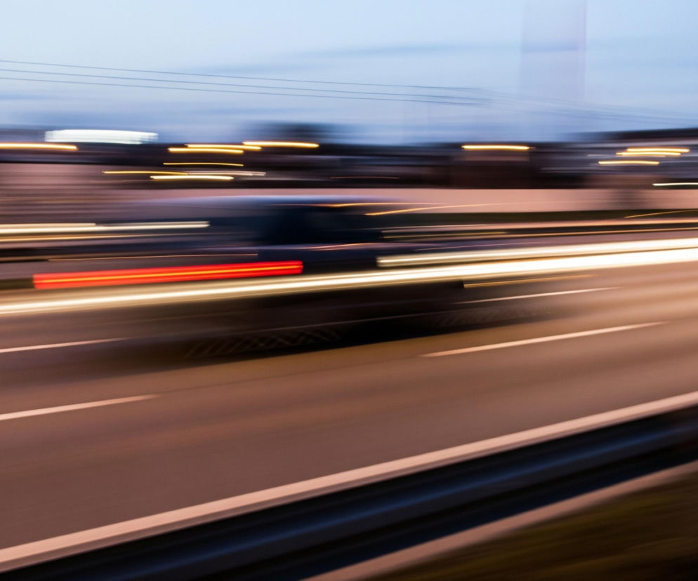 Rozmazana autostrada biegnie ukośnie przez środek kadru, z wyraźnymi białymi liniami pasów ruchu rozciągającymi się horyzontalnie. Czarne, zamazane sylwetki samochodów i jaskrawe, rozmyte smugi świateł – białe i żółte z przodu oraz czerwone z tyłu – ciągną się wzdłuż drogi, tworząc wrażenie ruchu. Górna część obrazu przedstawia rozmyte tło zabudowań i jasne, żółtawe punkty świateł miejskich, a nad nimi rozciąga się zachmurzone, niebiesko-szare niebo.