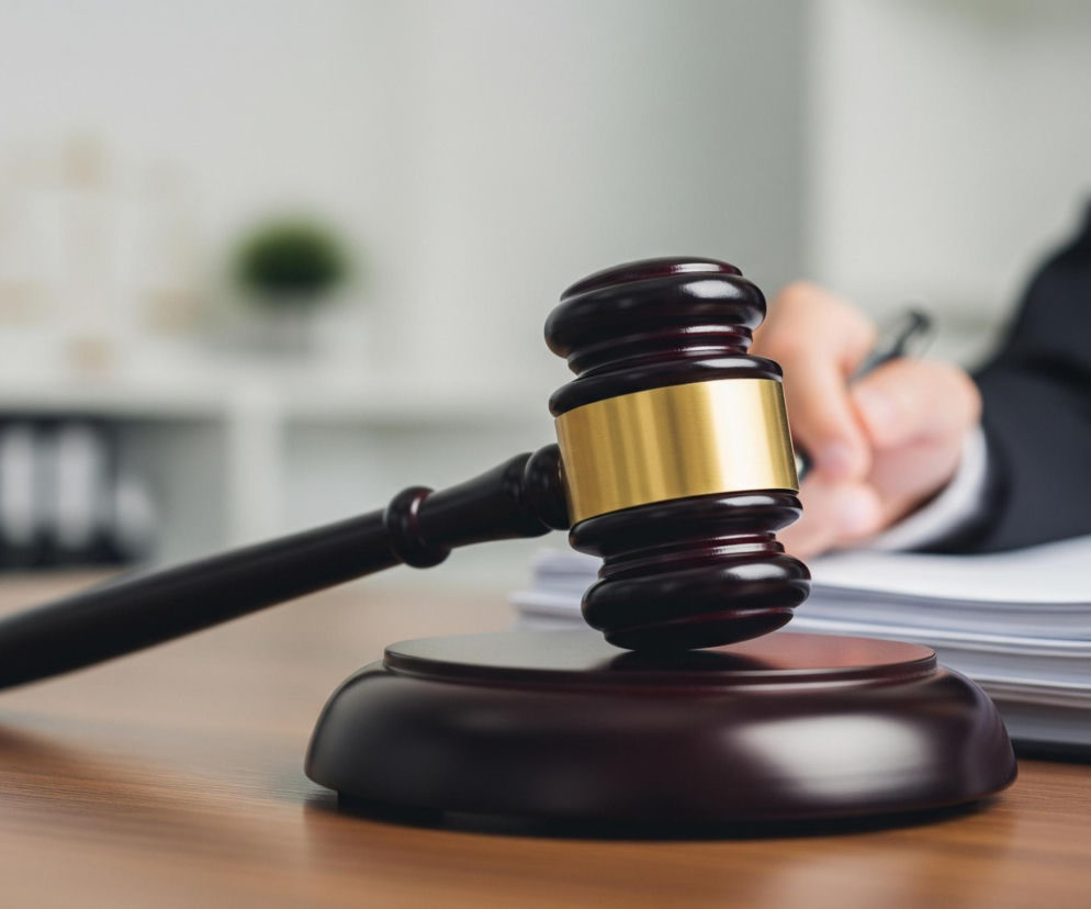 Na pierwszym planie, z lewej strony, na drewnianym blacie biurka leży ciemnobrązowy młotek sędziowski z błyszczącym złotym pierścieniem na głowicy, oparty o pasującą, okrągłą podstawę. Za młotkiem, po prawej stronie, widać stos dokumentów w czarnej teczce, na których spoczywa dłoń osoby w ciemnej marynarce, trzymającej długopis i piszącej. Tło jest rozmyte, z widocznymi jasnymi półkami i ciemnymi, prostokątnymi przedmiotami, a także zieloną rośliną doniczkową.