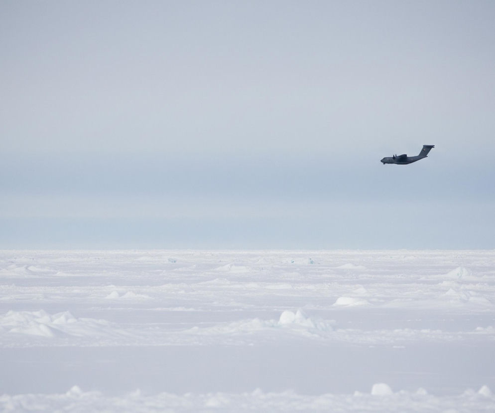 Ciemny samolot z dwoma silnikami pod skrzydłami, lecący w prawą stronę na tle jasnego, szaro-niebieskiego nieba. Poniżej samolotu rozciąga się rozległy, płaski krajobraz pokryty śniegiem i lodem, z licznymi pofalowanymi wzniesieniami i nierównościami, tworzącymi białą powierzchnię. Linia horyzontu jest wyraźna, oddzielając jaśniejsze niebo od bieli terenu.