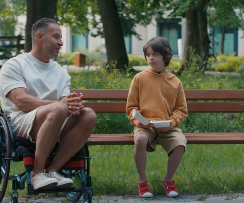 Mężczyzna na wózku inwalidzkim, Sebastian (Marek Krupski), rozmawia z chłopcem, Mateuszkiem (Filip Kowalewicz), siedzącym na ławce. Chłopiec trzyma pudełko z jedzeniem, wyraźnie smutny. Scena z serialu „Barwy szczęścia”, którą przeczytasz na portalu Super Seriale.