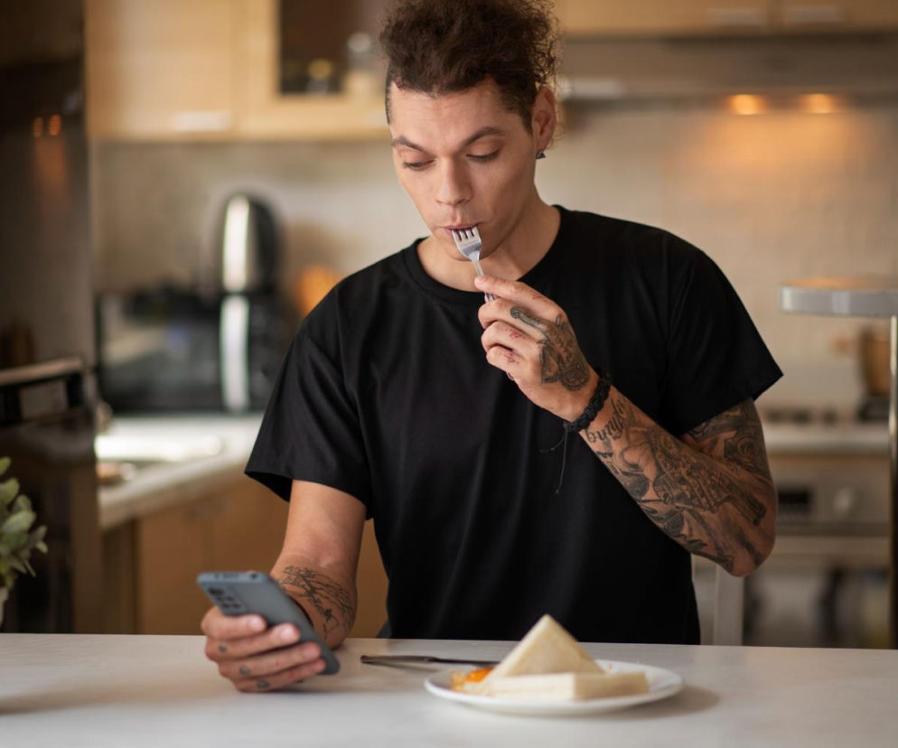 Mężczyzna w czarnym T-shircie je posiłek, trzymając widelec z jedzeniem blisko ust, jednocześnie patrząc w telefon komórkowy. Na talerzu widoczne są kanapki. Brak skupienia na jedzeniu to jeden z błędów prowadzących do problemów trawiennych, o czym przeczytasz na Poradnik Zdrowie.