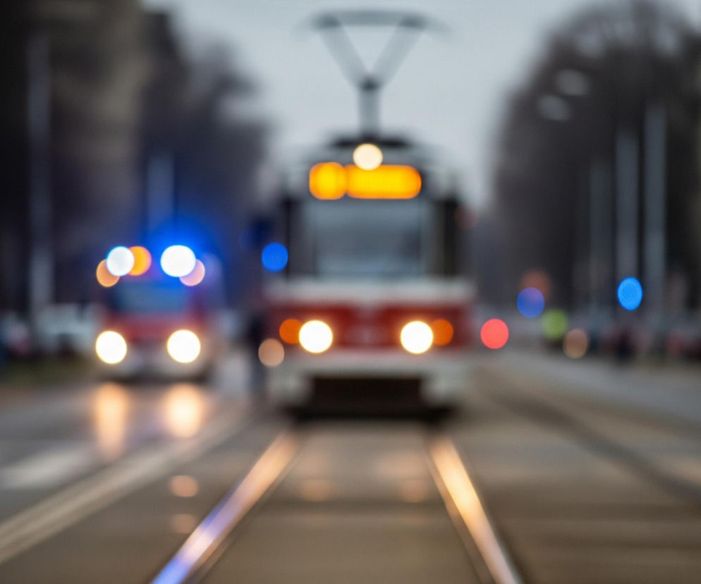 Rozmyte zdjęcie przedstawia ulicę z torami tramwajowymi biegnącymi przez środek. Na torach widoczny jest tramwaj, a obok niego, po lewej stronie, jedzie pojazd uprzywilejowany z migającymi niebieskimi światłami. Reflektory obu pojazdów są mocno rozświetlone, tworząc jasne, okrągłe punkty światła w przeważnie ciemnym otoczeniu. Po bokach ulicy znajdują się zaparkowane samochody oraz zarysy drzew i budynków w tle, wszystko jest rozmazane, co tworzy efekt bokeh.