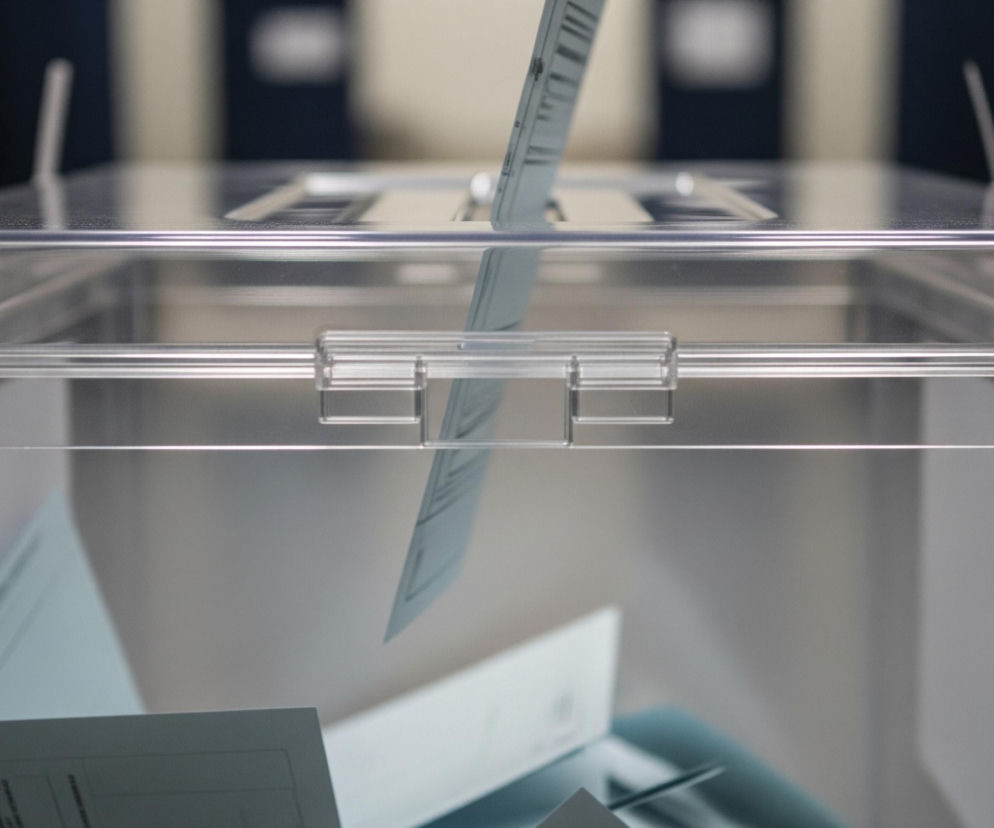 Na pierwszym planie widoczna jest przezroczysta, prostopadłościenna urna wykonana z akrylu. Z góry do szczeliny w jej wieku jest wkładana kartka, lekko zagięta, widoczna w górnej części kadru. Wewnątrz urny leży już kilka innych, jasnych kartek, częściowo zakrywających się nawzajem. Tło za urną jest rozmyte, z widocznymi pionowymi pasami w odcieniach ciemnego granatu i jasnego beżu, tworzącymi rytmiczny wzór.