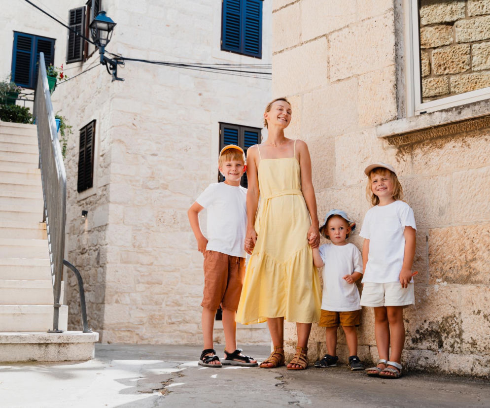 Uśmiechnięta mama w żółtej sukience spaceruje z trzema synami w jasnych ubraniach po kamiennej uliczce śródziemnomorskiego miasteczka. Dzieci noszą letnie czapki i sandały. Artykuły o udanych wakacjach z rodziną znajdziesz na naszym portalu.