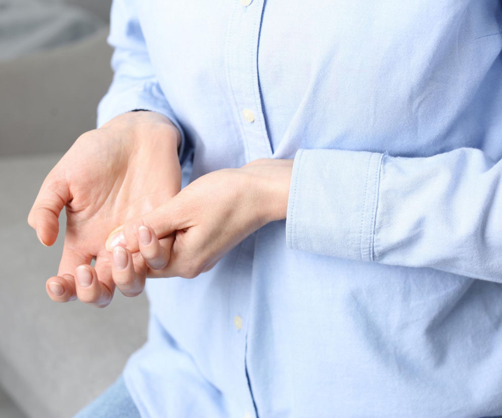 Kobieta w błękitnej koszuli trzymająca się za drżącą dłoń, symbolizująca obawę o drżenie rąk po kawie lub w spoczynku, co może być objawem choroby Parkinsona. Więcej informacji na ten temat znajdziesz w artykule na Poradnik Zdrowie.