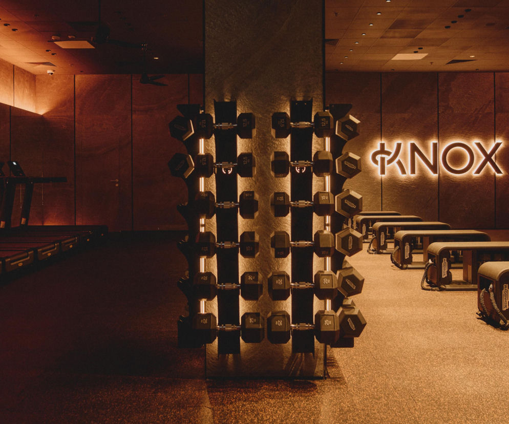 Wnętrze butikowego studia fitness KNOX z rzędami bieżni po lewej i strefą do treningu siłowego z ławkami i hantlami po prawej, oświetlone ciepłym, pomarańczowym światłem tworzącym immersyjną atmosferę treningu. Na tylnej ścianie widoczny podświetlany neon z logo KNOX. Więcej o nowej metodzie Run & Lift przeczytasz na naszym portalu.