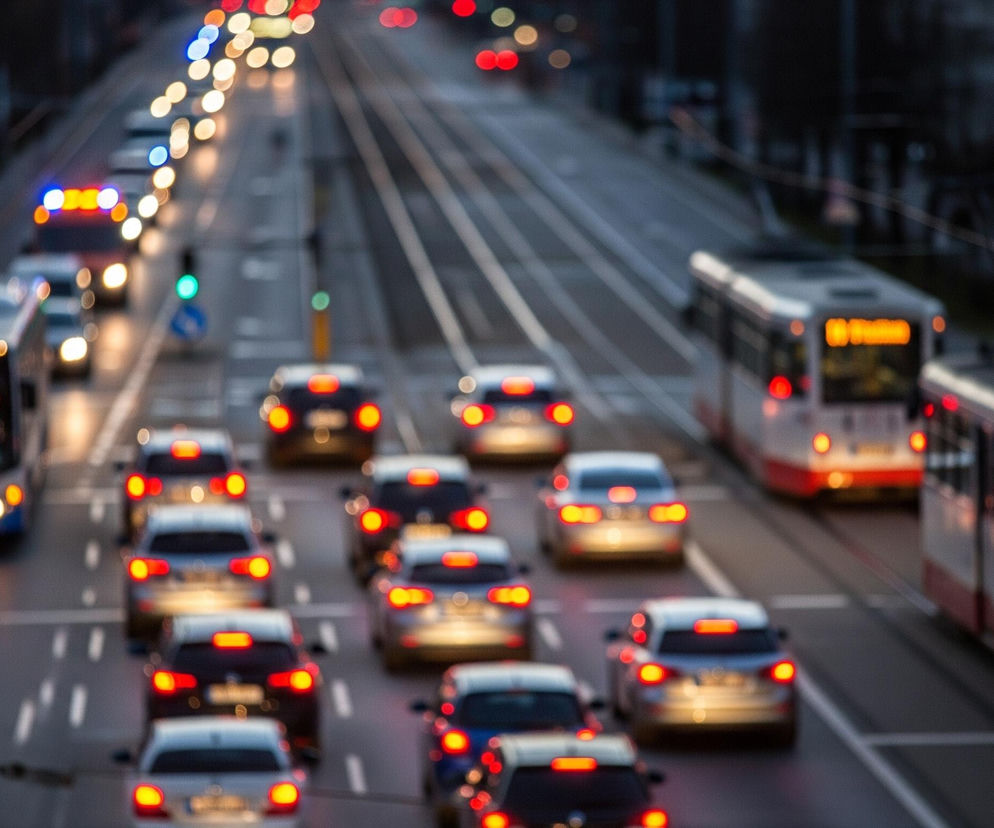 Na zdjęciu widoczna jest miejska ulica o zmierzchu lub w nocy, z licznym ruchem pojazdów. W centralnej części obrazu znajduje się wiele samochodów i autobusów, z włączonymi światłami przednimi i tylnymi, tworzącymi świetliste, rozmyte plamy. Po prawej stronie biegną tory tramwajowe, na których widoczne są dwa tramwaje – jeden bliżej, biało-czerwony, a drugi dalej, biało-pomarańczowy, oba z włączonymi światłami i rozświetlonym wnętrzem. Cała scena jest rozmyta, co nadaje jej charakterystyczny efekt bokeh, a światła uliczne i pojazdów dominują w kolorach czerwieni, pomarańczy, żółci i błękitu, kontrastując z ciemnym tłem miasta.