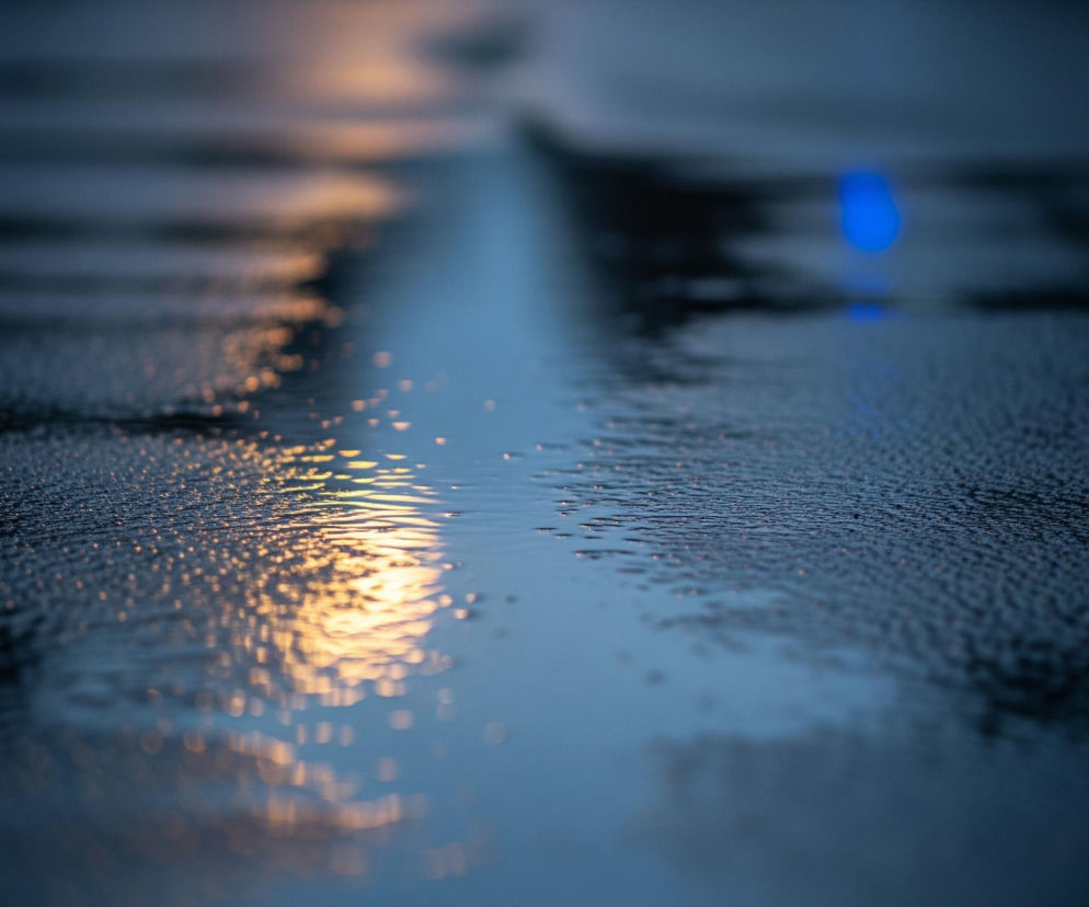 Na pierwszym planie widoczna jest mokra powierzchnia, prawdopodobnie asfaltu lub betonu, pokryta drobnymi kroplami wody, z których odbijają się rozproszone, pomarańczowe światła po lewej stronie. Przez środek obrazu, wzdłuż jego długości, biegnie pas bardziej intensywnych odbić światła, tworząc wrażenie mokrej smugi lub kałuży. W tle, po prawej stronie, widać rozmyte, niebieskie światło, odbijające się również od powierzchni. Całość utrzymana jest w ciemnych, chłodnych barwach, z dominującym granatem i odcieniami szarości.