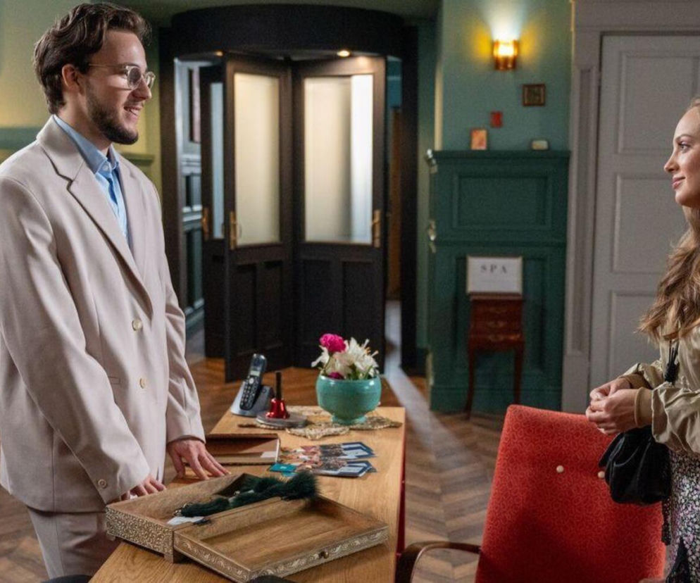 Oliwka (Wiktoria Gąsiewska) i Justin (Jasper Sołtysiewicz) w hotelu Stańskich, patrzący na siebie z uśmiechem. Oliwka w beżowej kurtce i wzorzystej spódnicy, Justin w beżowym garniturze i niebieskiej koszuli. Scena z serialu Barwy szczęścia dostępnego na Super Seriale.