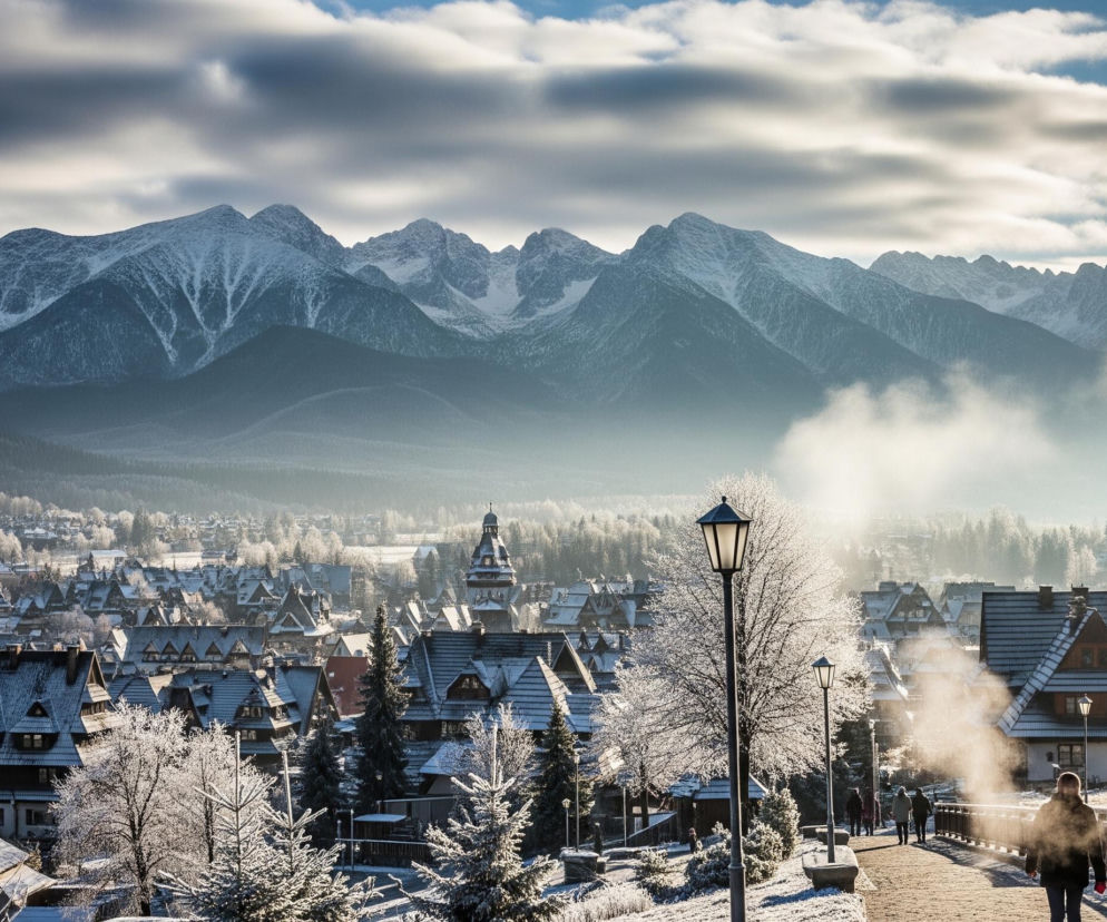 Widok zimowy przedstawia zaśnieżone szczyty górskie na horyzoncie, z pasmami chmur częściowo zakrywającymi błękitne niebo. Poniżej rozciąga się miejski krajobraz z licznymi budynkami o spadzistych, pokrytych śniegiem dachach, często z elementami architektonicznymi przypominającymi wieże i kopuły, niektóre z nich posiadają charakterystyczne jaskółki. W centrum kadru na pierwszym planie znajduje się metalowa latarnia uliczna, a obok niej białe, oszronione drzewo, pod którym rosną mniejsze, również oszronione krzewy. Po prawej stronie widoczna jest kamienista ścieżka lub ulica, po której poruszają się pojedyncze postaci, z których unoszą się opary pary, sugerujące niską temperaturę.