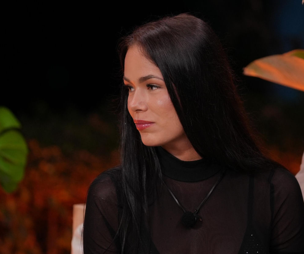 Magda z Hotelu Paradise na zbliżeniu, z długimi ciemnymi włosami i w przezroczystej czarnej bluzce, podczas odcinka z tajemniczą wiadomością od Rajskiego Boga. W tle widać ciemność i liść egzotycznej rośliny, co podkreśla scenerię reality show. Więcej o losach Magdy przeczytasz na Super Seriale.