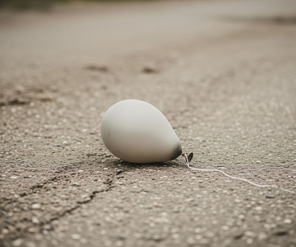 Pośrodku kadru, na popękanej nawierzchni asfaltowej, leży szary, owalny balon. Ma gładką, matową powierzchnię, odbijającą delikatnie światło. Z jego dolnej części wystaje zawiązany sznurek, który ciągnie się w prawą stronę, biegnąc wzdłuż pęknięcia w nawierzchni. Tło jest rozmyte, ukazując jaśniejszą, jednolitą przestrzeń drogi.