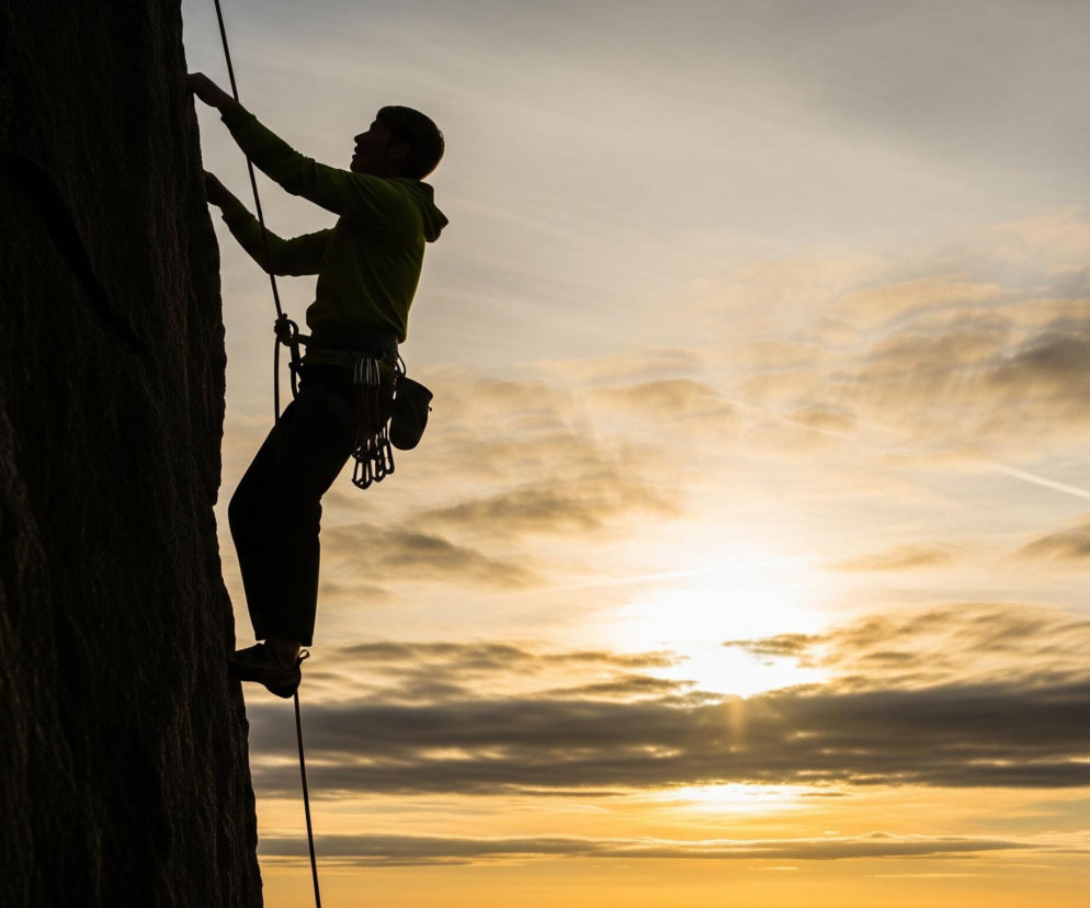 Wysoki, ciemny profil wspinacza, zarysowany na tle zachodzącego słońca, dominuje lewą stronę kadru. Mężczyzna w zielonej bluzie z kapturem i spodniach trzyma się obiema rękami ciemnej, skalistej ściany, a jego prawa stopa opiera się na niewidocznym występie. Do jego uprzęży przyczepiony jest sprzęt wspinaczkowy, a lina biegnie w dół poza kadr. Horyzont na tle pomarańczowo-żółtego nieba, z rozproszonymi chmurami, znajduje się w prawym dolnym rogu. Promienie słońca przebijają się przez chmury, tworząc złocisty blask, a na niebie widoczne są również smugi kondensacyjne.