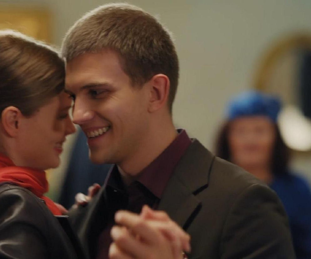 Młoda kobieta w czarnej kurtce i czerwonym szaliku tańczy z uśmiechniętym mężczyzną w garniturze. Para patrzy na siebie, a w tle widać rozmazane sylwetki innych gości. To romantyczna scena z serialu M jak miłość, o której więcej dowiesz się na Super Seriale.