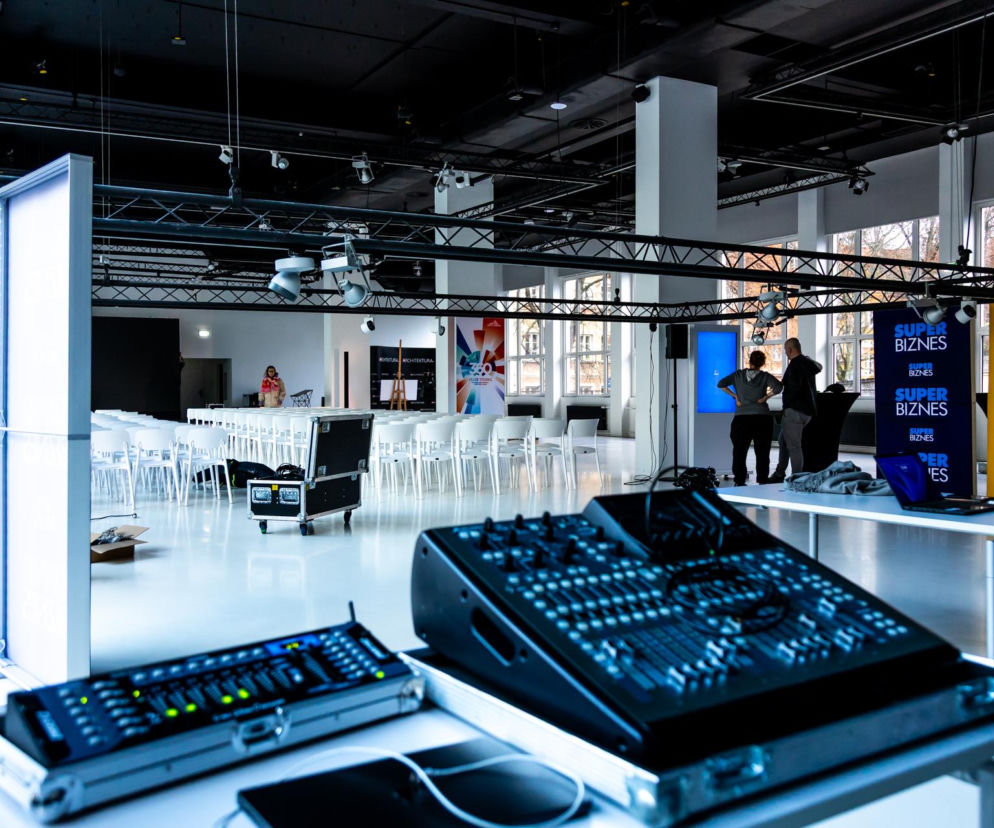 Sala konferencyjna przygotowywana na galę konkursu Życie w Architekturze, z rzędami białych krzeseł, sceną z oświetleniem i stanowiskiem technicznym. Widoczne banery Architektura Murator Plus i logotypy Murator. Scena czeka na finał 10. edycji.