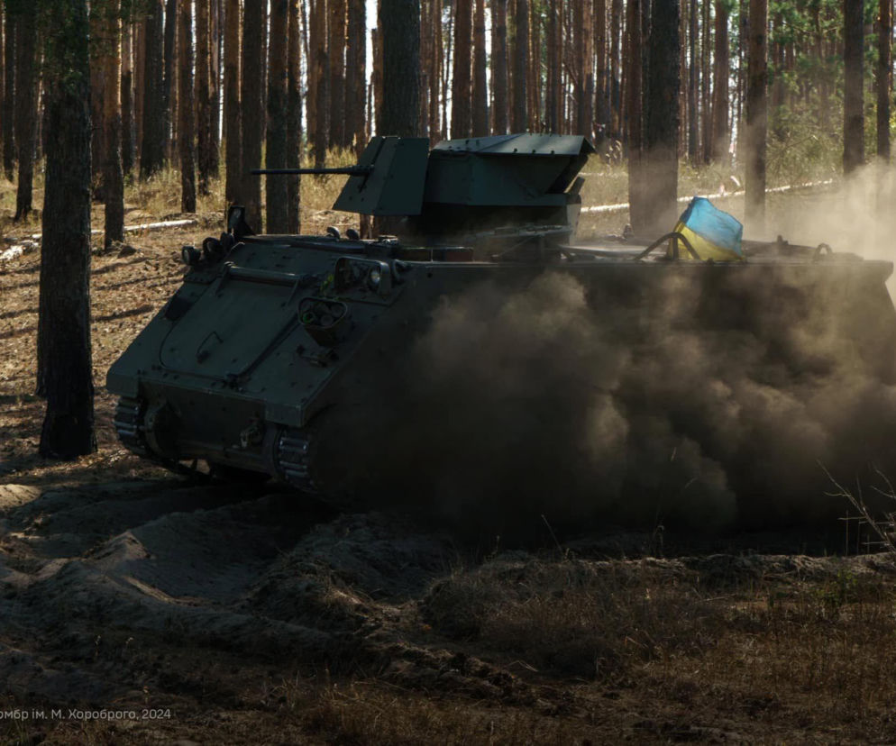Transporter opancerzony M113 w zielonym kamuflażu, z flagą Ukrainy, porusza się w piaszczystym terenie leśnym, unosząc tumany kurzu, co obrazuje militarne wsparcie Norwegii dla Ukrainy, o którym możesz przeczytać na naszym portalu.