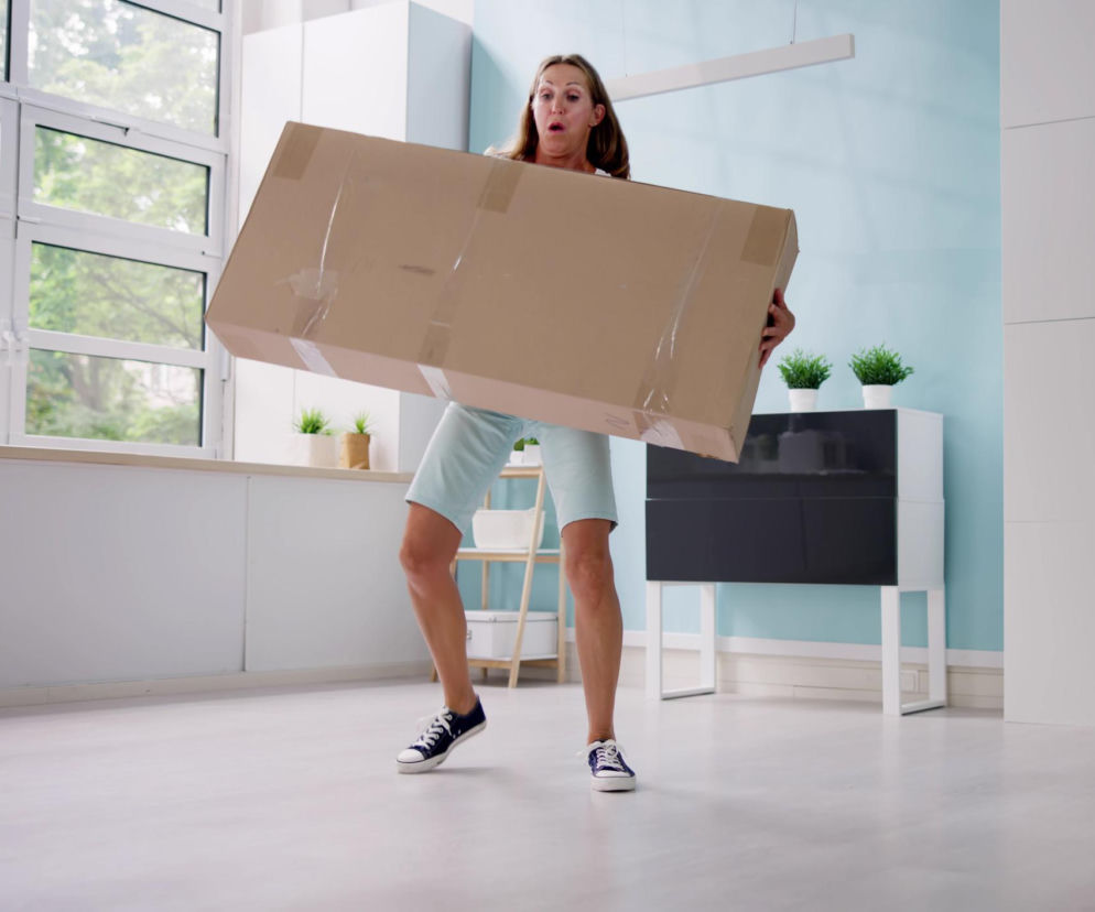 Kobieta w sportowym stroju, ze zdziwioną miną, próbuje podnieść dużą, ciężką paczkę. To często popełniany błąd przez osoby z nadciśnieniem, które wstrzymują oddech przy wysiłku, co może prowadzić do zawału lub udaru, jak informuje Poradnik Zdrowie.