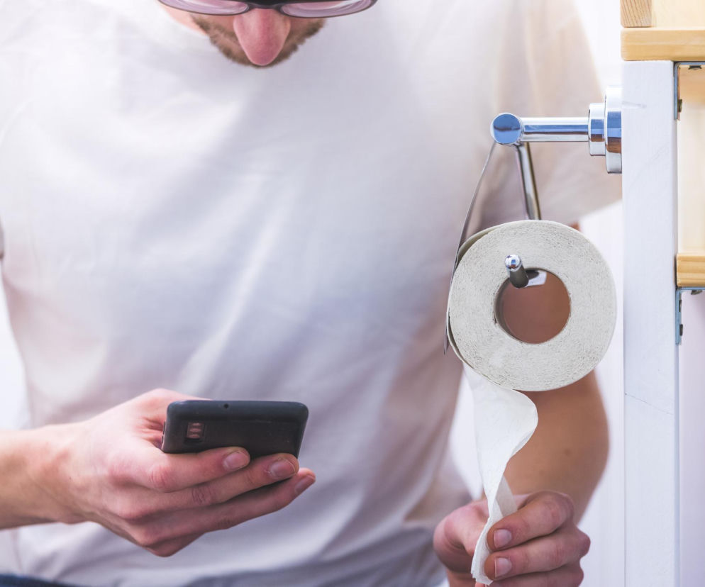 Mężczyzna w okularach i białej koszulce używający smartfona w toalecie, obok niego wieszak z papierem toaletowym. Scena przedstawia powszechny nawyk, który, jak czytamy na Poradnik Zdrowie, zwiększa ryzyko hemoroidów i bólu pleców.
