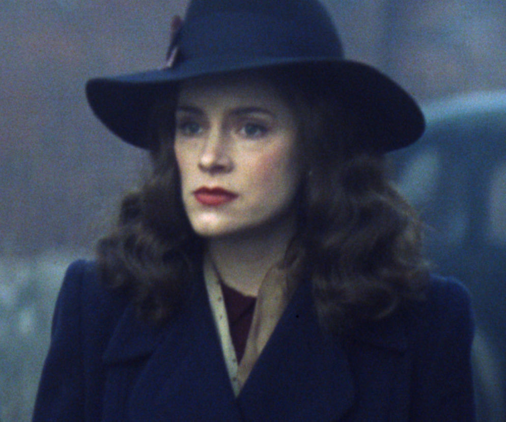 Kobieta w granatowym kapeluszu i płaszczu z falowanymi włosami i czerwonymi ustami. Jej spojrzenie jest intensywne, a wyraz twarzy sugeruje tajemniczość i powagę. Zdjęcie w stylu retro pasuje do tematyki powrotu kultowych Super Seriale Peaky Blinders.