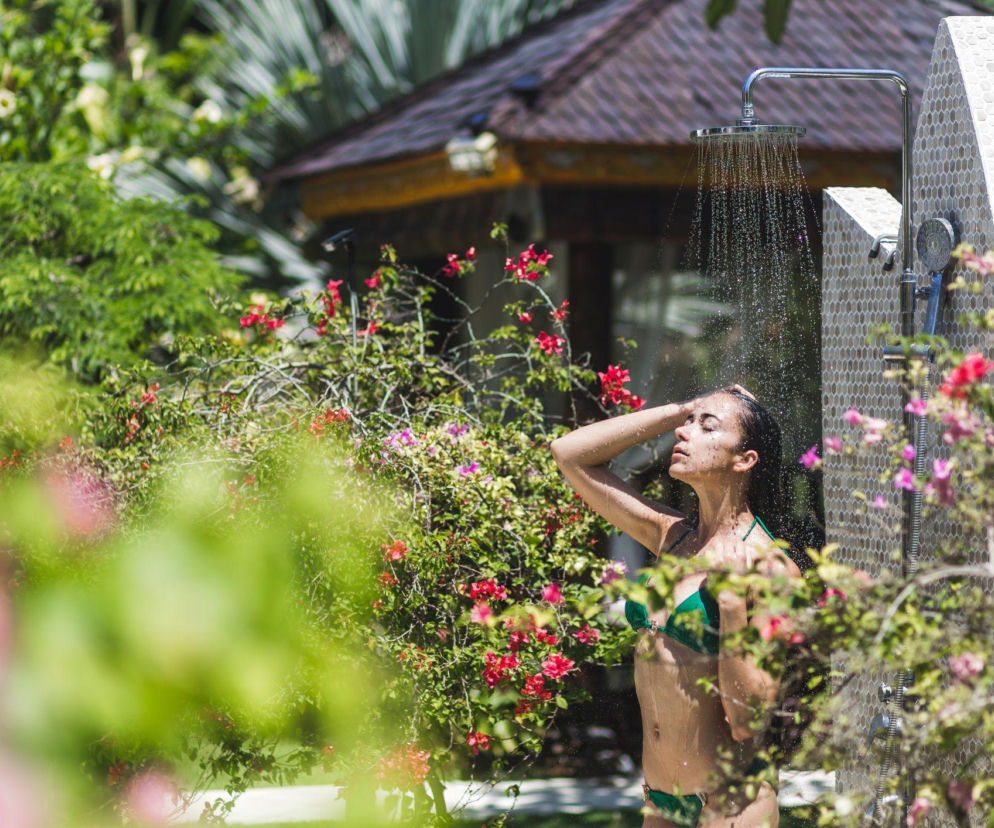 Kobieta w zielonym bikini odświeżająca się pod deszczownicą prysznica ogrodowego, otoczonego bujną zielenią i kwitnącymi krzewami. Idealny sposób na ochłodę w upalne dni. Więcej o prysznicach ogrodowych przeczytasz na naszym portalu.