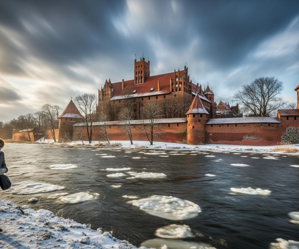 Kobieta w szarym płaszczu i zimowej czapce z futrem, z rozwianym szalikiem, idzie po pokrytym śniegiem brzegu rzeki, która płynie od lewej do prawej, usiana licznymi kawałkami lodu. Na prawym brzegu, w tle, widoczny jest masywny ceglany zamek z czerwonymi dachami, składający się z wielu wież i budynków, częściowo otoczony murem obronnym, przed którym rosną nagie drzewa. Niebo jest dynamiczne, z rozciągniętymi, rozmytymi chmurami w odcieniach szarości i błękitu, z jaśniejszym, złotawym blaskiem słońca po lewej stronie.