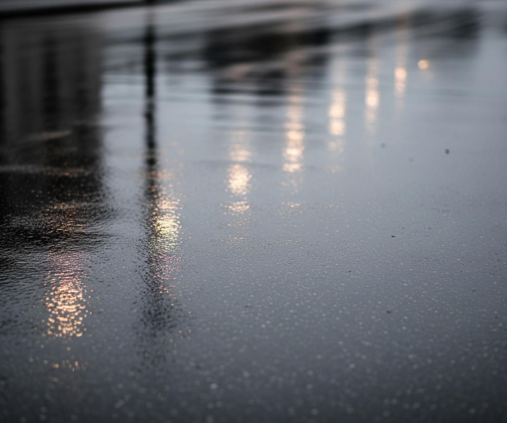 Ulica lub plac pokryty jest licznymi kroplami wody, odbijającymi jasne, rozmyte światła w oddali. Dominują ciemne odcienie szarości i czerni na mokrej powierzchni, która jest głównym elementem kadru. W górnej części widoczne są niewyraźne, ciemne kształty budynków oraz pojedynczy, pionowy, ciemny słup, którego odbicie również jest widoczne na nawierzchni. Po prawej stronie szereg jasnych, pomarańczowo-żółtych plam światła, prawdopodobnie lamp ulicznych, rozciąga się w głąb obrazu, z odbiciami również na mokrej powierzchni.