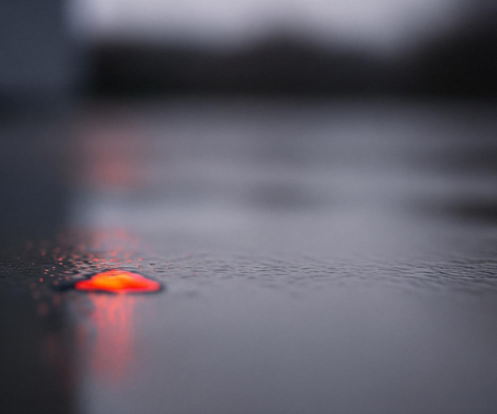 Czerwony, rozmyty obiekt o nieregularnym kształcie leży na mokrej, ciemnej powierzchni, która odbija jego jaskrawy kolor. Powierzchnia, przypominająca asfalt lub beton, jest wilgotna, z niewielkimi fałdami lub nierównościami, widocznymi szczególnie na horyzoncie. Tło jest bardzo rozmyte, utrzymane w ciemnych, szarych i czarnych tonach, co potęguje kontrast z wyraźnym, czerwonym obiektem na pierwszym planie.