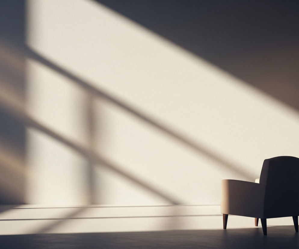 Beżowy fotel stoi w prawym dolnym rogu pomieszczenia, oświetlony częściowo promieniami słońca. Duża część tła to jasna ściana, na którą padają ukośne pasma światła i cienia, tworzące geometryczne wzory. Podłoga jest ciemniejsza, również z widocznymi promieniami słonecznymi.