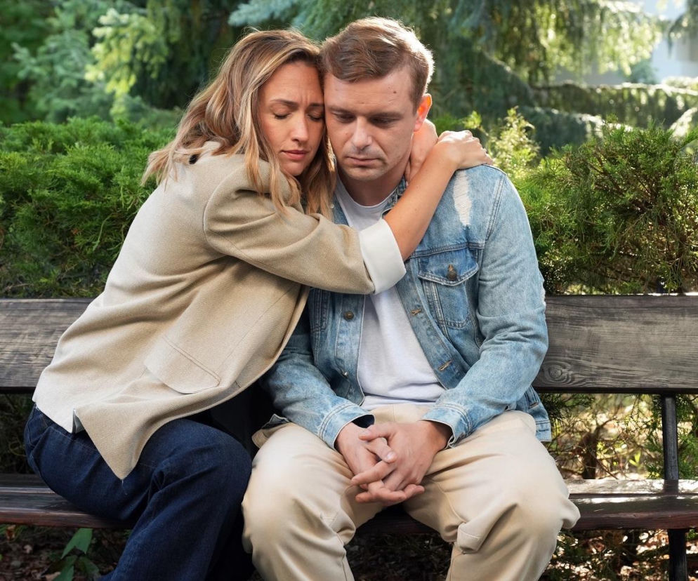Asia (Anna Gzyra) w beżowej marynarce obejmuje siedzącego obok niej Huberta (Marek Molak) w jeansowej kurtce, oboje z zamkniętymi oczami, smutni, na tle zielonych krzewów. To kadr z serialu Barwy szczęścia, a więcej o tym wątku przeczytasz na stronie Super Seriale.