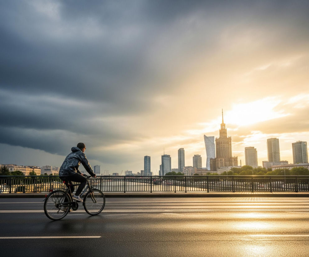Mężczyzna w ciemnej kurtce z kapturem i czarnych spodniach jedzie na rowerze w prawą stronę, widoczny od tyłu, po mokrej, ciemnoszarej nawierzchni z białymi liniami. Po prawej stronie widoczna jest latarnia uliczna. W tle rozciąga się miejski krajobraz z wieloma wysokimi budynkami, w tym Pałacem Kultury i Nauki, z rzeką i barierkami mostu poniżej. Niebo jest dramatycznie podzielone: po lewej stronie ciemne, burzowe chmury, a po prawej jasne, złote światło słoneczne, rzucające ciepłe refleksy na wodę i budynki.