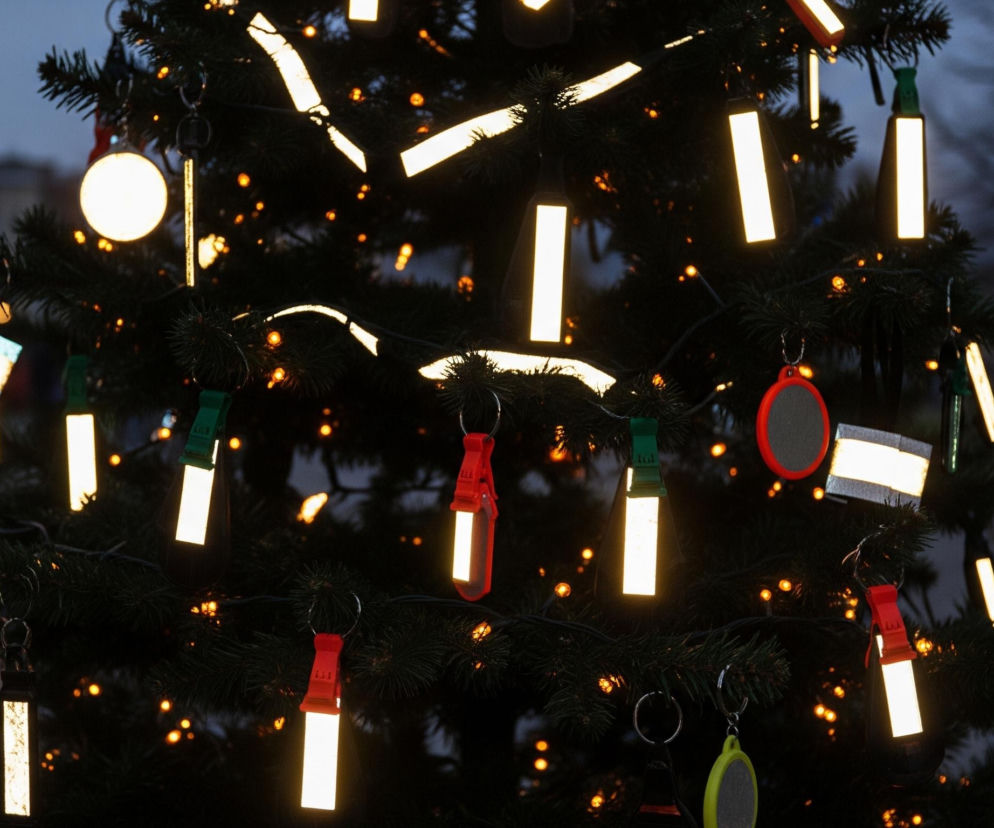 Na pierwszym planie widoczna jest choinka udekorowana licznymi ozdobami odblaskowymi w różnych kształtach, takich jak prostokąty, paski i okręgi. Ozdoby te, w przeważającej części prostokątne, intensywnie odbijają światło, świecąc jasnożółtym kolorem na tle ciemnozielonych gałęzi. Pomiędzy odblaskowymi elementami widać mniejsze, ciepłe, żółtopomarańczowe światełka choinkowe. W tle rozmazane sylwetki budynków i drzew oraz pojedyncze, rozmyte punkty światła w dali tworzą wieczorny, miejski krajobraz.