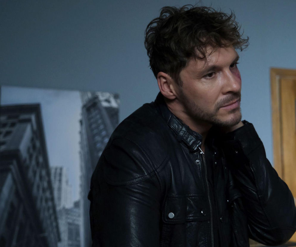 Aktor Mikołaj Roznerski jako Marcin Chodakowski z serialu M jak miłość z widoczną raną na policzku, ubrany w czarną skórzaną kurtkę, spogląda w prawą stronę kadru. Obraz doskonale ilustruje kryzys emocjonalny bohatera, o którym przeczytasz więcej na Super Seriale.