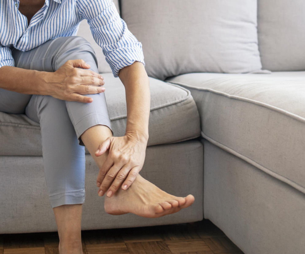 Osoba siedząca na kanapie chwytająca się za kostkę, co sugeruje ból lub dyskomfort stawów. To wizualizacja pierwszych sygnałów zapalenia stawów, o których przeczytasz więcej na Poradnik Zdrowie.