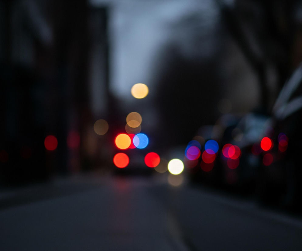 Ulica nocą rozmywa się w nieostre plamy świateł. W centrum obrazu dominują jasne, okrągłe światła: jedno niebieskie, jedno pomarańczowe i wiele czerwonych, ułożone w chaotyczną kompozycję. Po bokach widać zarys ciemnych budynków lub drzew, również nieostrych, a na pierwszym planie znajduje się ciemny asfalt ulicy z delikatnym jasnym paskiem, prawdopodobnie linią jezdni, prowadzącym w głąb obrazu.
