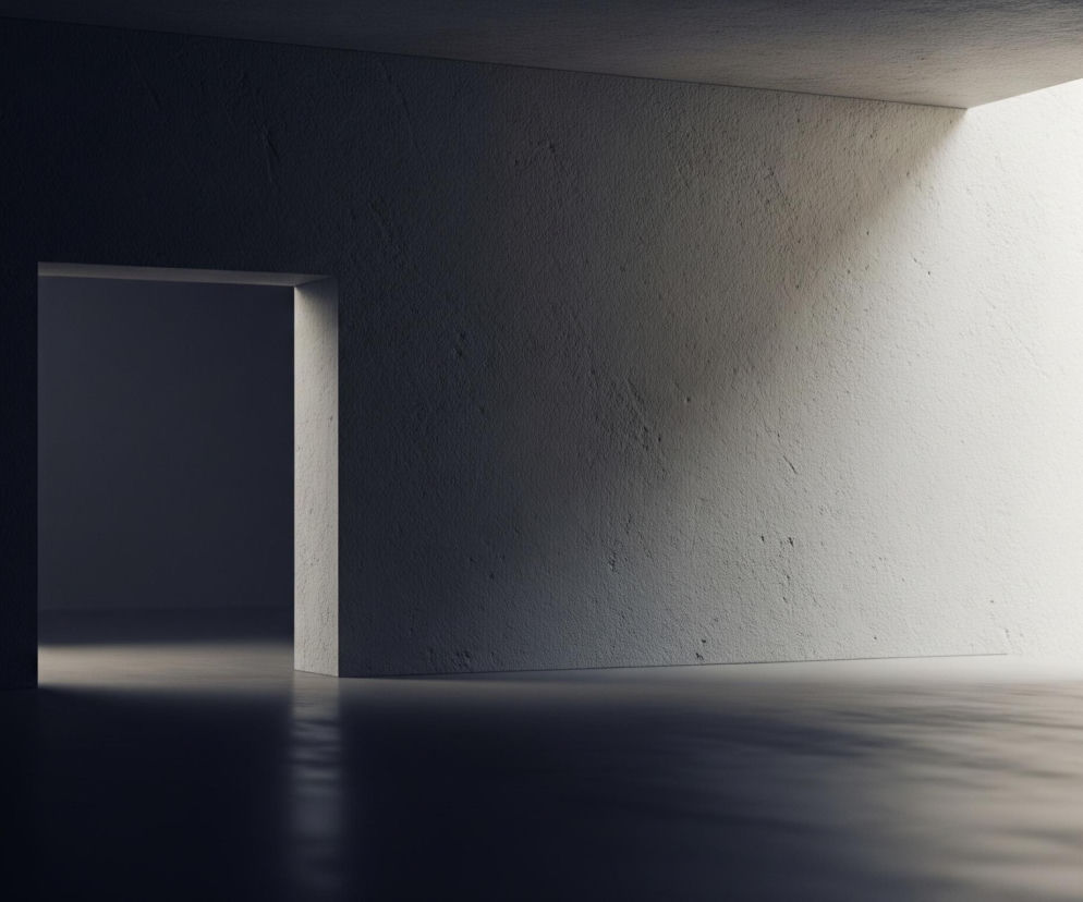 Wnętrze pomieszczenia z drzwiami po lewej stronie i jasnym przejściem po prawej. Dominują odcienie szarości i bieli; lewa ściana i sufit są w ciemnym cieniu, z widoczną fakturą, natomiast prawa ściana jest intensywnie oświetlona, co tworzy silny kontrast. Jasne światło padające z prawej strony oświetla również podłogę, tworząc na niej refleksy i cienie, szczególnie w kierunku drzwi, które prowadzą do ciemniejszego, głębiej położonego pomieszczenia.