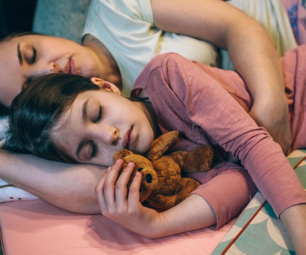 Matka z córką śpiące obok siebie na łóżku. Dziewczynka w różowej bluzce z piegami na twarzy przytula pluszowego misia, a jej mama leży obok. Zdrowy sen jest kluczowy, a więcej na ten temat przeczytasz na portalu Poradnik Zdrowie.