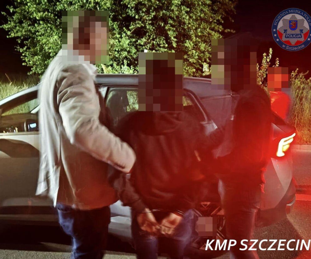 Policjant prowadzi skutego kajdankami mężczyznę, podejrzanego o zabójstwo w Szczecinie, w kierunku radiowozu. Scena dzieje się w nocy, w tle widać drzewa i logo Komendy Miejskiej Policji w Szczecinie. Więcej informacji o tej sprawie przeczytasz na naszym portalu.