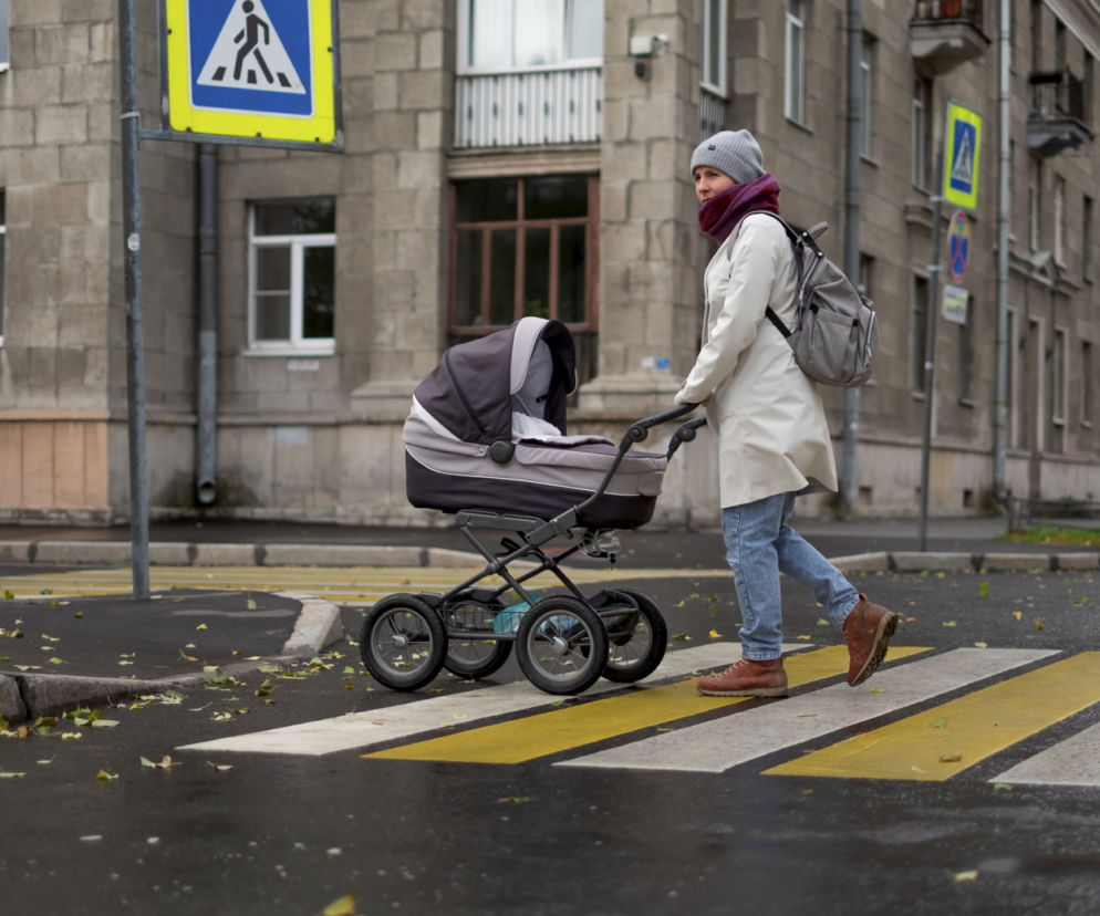 Kobieta prowadzi podwójny wózek bliźniaczy przez przejście dla pieszych. O wyborze modelu przeczytasz na SE.