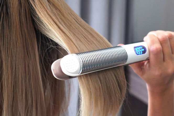 Dłoń osoby przesuwającej prostownicę z wyświetlaczem po pasmie blond włosów. O stylizacji pasm przeczytasz na PZ Uroda.