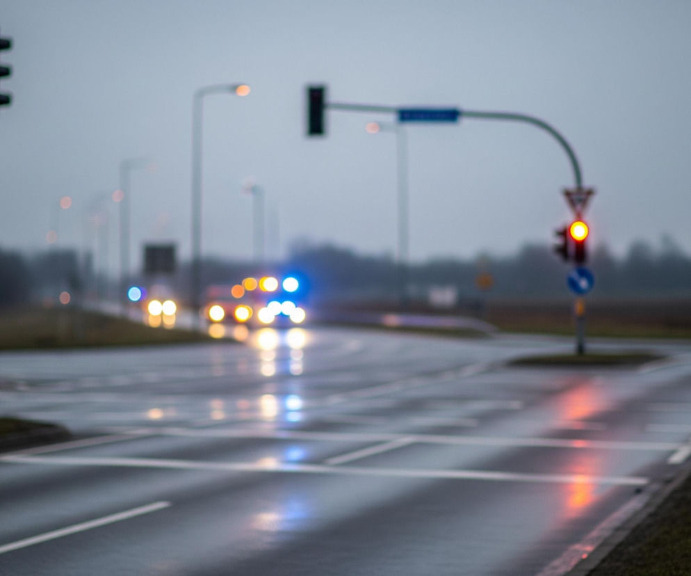 Rozmyty widok skrzyżowania dróg w pochmurny dzień, prawdopodobnie po deszczu. Na pierwszym planie po lewej stronie znajduje się słup z dwoma sygnalizatorami świetlnymi, jeden nad drugim, oba wydają się być wygaszone. Dalej, po prawej stronie, widać drugi sygnalizator świetlny z wyraźnie świecącym czerwonym światłem oraz drogowskaz w kształcie trójkąta z czerwonym obramowaniem.

W tle widoczne są światła nadjeżdżających samochodów – jasne reflektory oraz niebieskie pulsujące światła uprzywilejowanych pojazdów, odbijające się na mokrej nawierzchni jezdni. Wzdłuż drogi rozmieszczone są latarnie uliczne, emitujące ciepłe, pomarańczowe światło. Otoczenie skrzyżowania stanowią niewyraźne zarysy drzew i otwartych przestrzeni, a niebo ma szary, jednolity odcień.