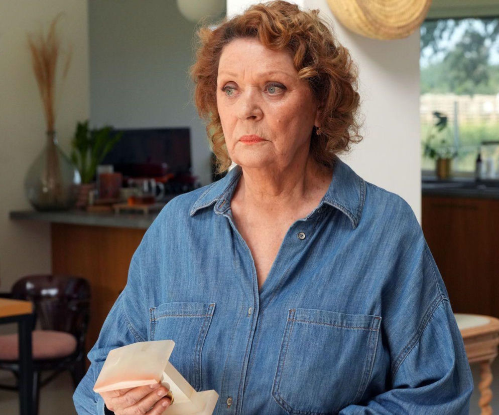 Starsza kobieta, Józefina (Elżbieta Jarosik), z rudymi, kręconymi włosami i zmartwionym wyrazem twarzy, w jeansowej koszuli, trzyma w dłoniach małe, beżowe pudełeczko. W tle widać fragmenty nowoczesnego wnętrza salonu lub kuchni. Zdjęcie ilustruje zmiany w życiu Józefiny w serialu Barwy szczęścia, o czym więcej przeczytasz na portalu Super Seriale.