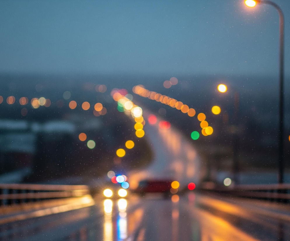 Długi, rozświetlony most rozciąga się w głąb obrazu, zajmując centralną część kadru. Nawierzchnia mostu jest mokra, co odbija światła samochodów i latarni, tworząc smugi w odcieniach pomarańczy, żółci, błękitu i czerwieni. Po prawej stronie, na pierwszym planie, wznosi się metalowy słup z jasno świecącą, pomarańczową lampą uliczną, a poniżej widoczna jest brązowa barierka mostu. W oddali, za mostem, rozciąga się panorama miasta z licznymi rozmytych, kolorowymi światłami. Cała scena spowita jest w odcieniach szarości i błękitu, sugerując zmierzch lub noc, a w powietrzu unosi się delikatny deszcz lub śnieg, widoczny jako drobne, jasne punkty.