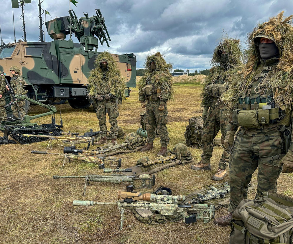 Żołnierze w mundurach maskujących i strojach ghillie prezentujący broń snajperską oraz sprzęt wojskowy, w tym pojazd opancerzony i robota saperskiego, na poligonie. Taka przygoda może czekać młodych podczas Wakacji z wojskiem, o których przeczytasz na Super Biznes.