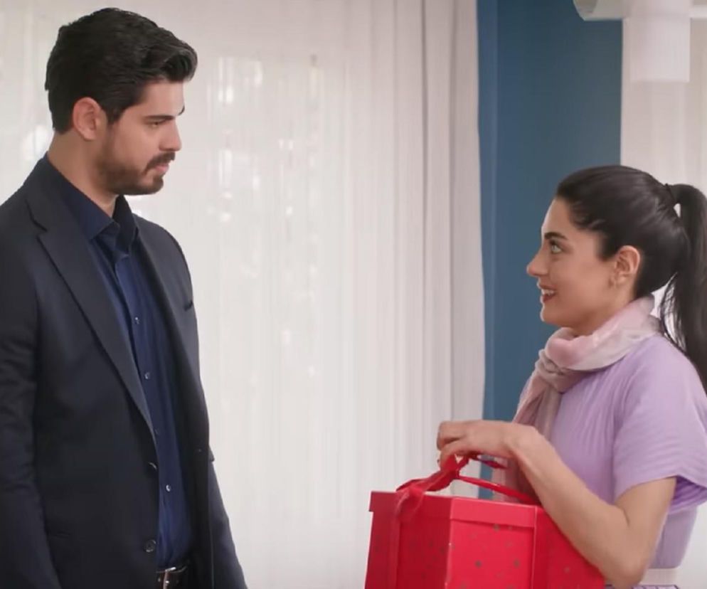 Cihan (Türkseve) w ciemnym garniturze i Hancer (Talya Çelebi) w fioletowej bluzce z różowym szalikiem, trzymająca czerwone pudełko prezentowe. Scena z serialu Panna młoda, w której Cihan wręcza żonie prezenty z Dubaju. Więcej o tym na Super Seriale.