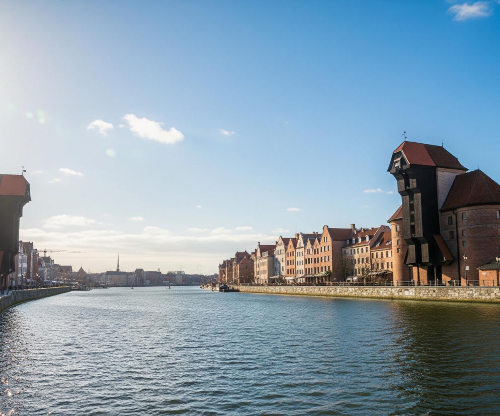 Rzekę płynącą przez miasto, otoczoną z obu stron zabudowaniami i nadrzecznymi promenadami. Po prawej stronie dominują kamienice o ceglanych i pastelowych fasadach, z widocznym masywnym drewnianym Żurawiem w centralnej części, który posiada czerwony spadzisty dach i ciemnobrązową konstrukcję. Na lewym brzegu również znajdują się budynki i drugi, mniejszy dźwig z czarnym drewnianym elementem. W dali, wzdłuż rzeki, rozciąga się panorama miasta z licznymi dachami, wieżami i spiczastym hełmem. Na pierwszym planie, po lewej stronie, spaceruje kilka osób po brukowanej ścieżce, oświetlonej przez intensywne słońce, którego światło odbija się również od falującej powierzchni wody. Błękitne niebo z kilkoma białymi chmurami rozjaśnia scenę.