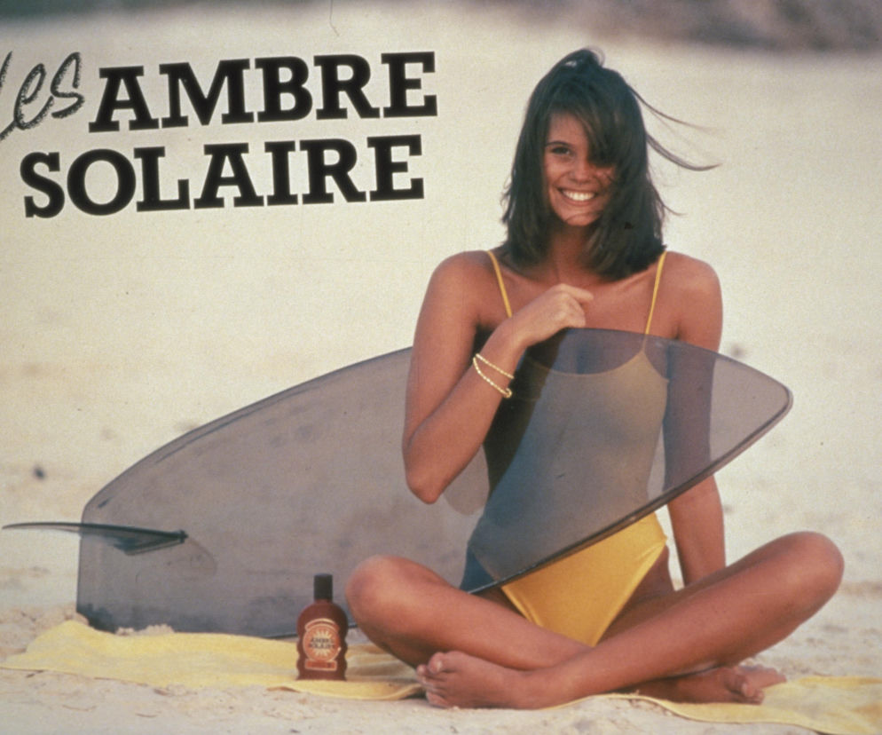 Uśmiechnięta Elle Macpherson na plaży z produktem Ambre Solaire. O historii marki przeczytasz na SE.