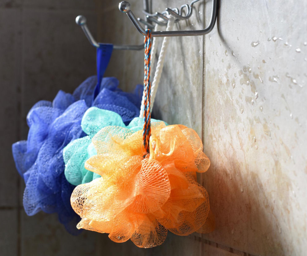 Trzy kolorowe gąbki myjki – niebieska, turkusowa i pomarańczowa – wiszą na metalowym haczyku pod prysznicem, ociekając wodą na mokre kafelki. Artykuł na Poradnik Zdrowie wyjaśnia, jak zły nawyk pod prysznicem, związany z gąbkami, może negatywnie wpływać na skórę.